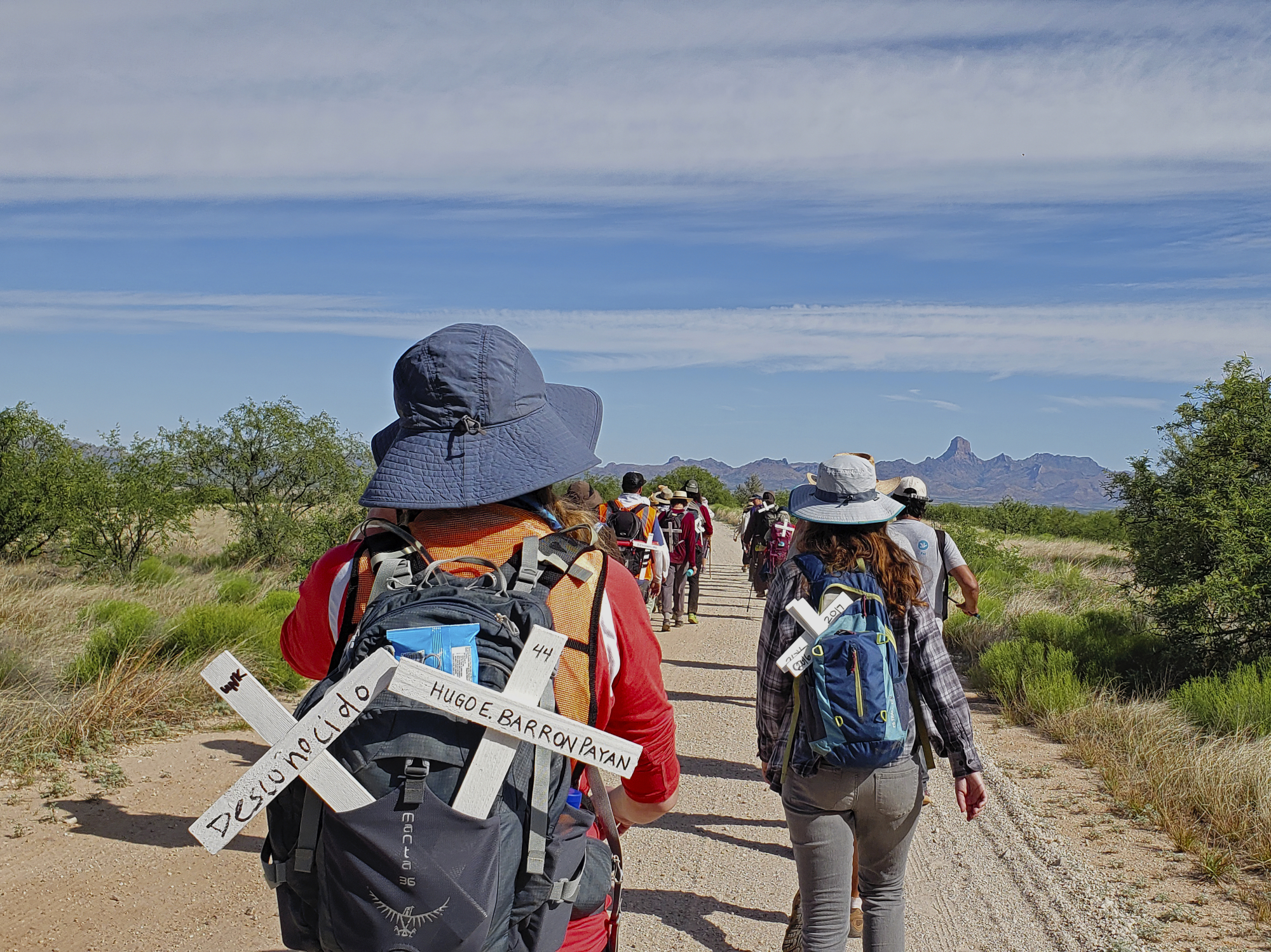 Un groupe de personnes marche sur un chemin de terre. Dans leurs sacs à dos, on trouve des croix avec des messages en espagnol.