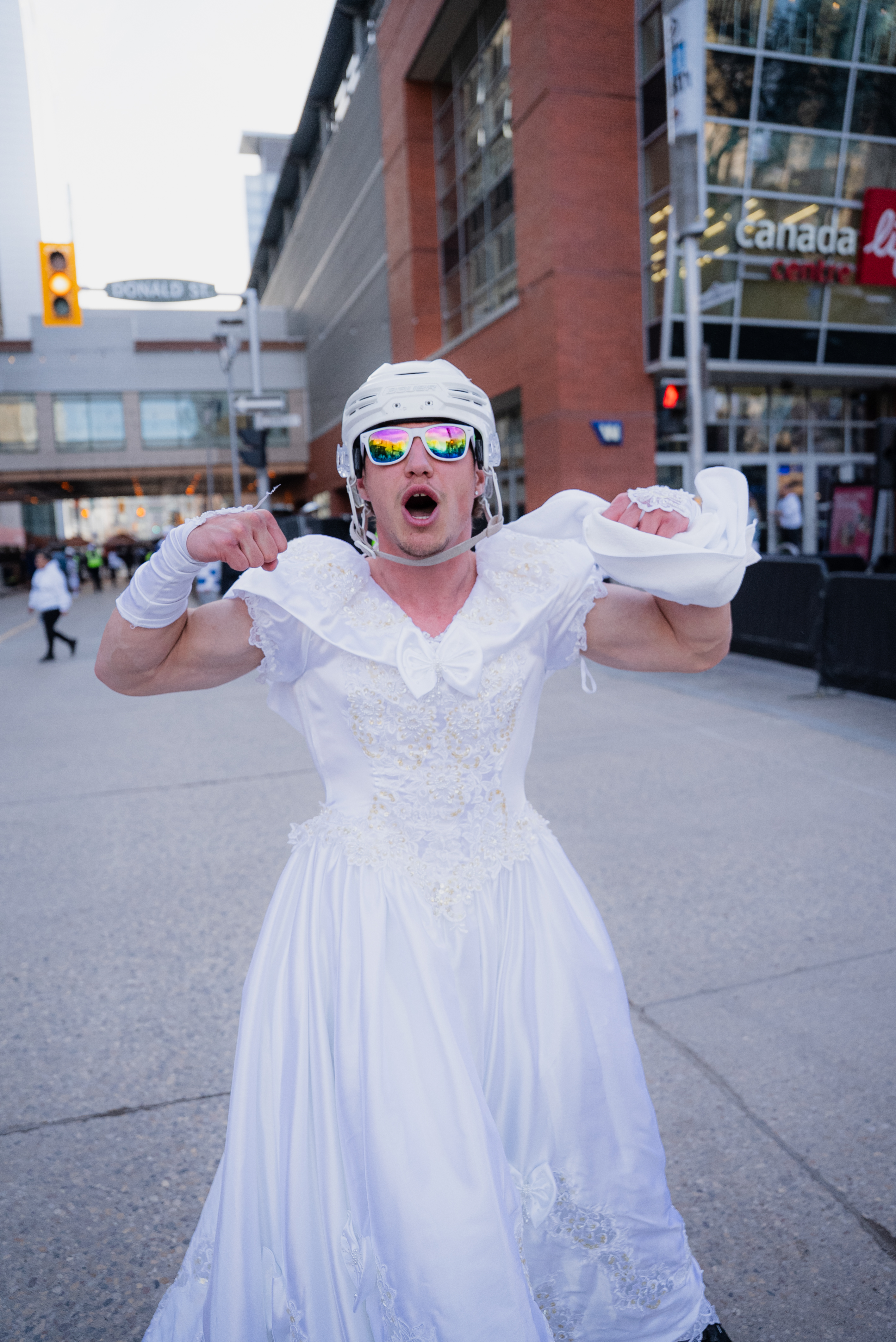 Man standing in downtown Winnipeg dressed in a white wedding dress, hockey helmet and sunglasses. More men in wedding dresses can be seen in the background.