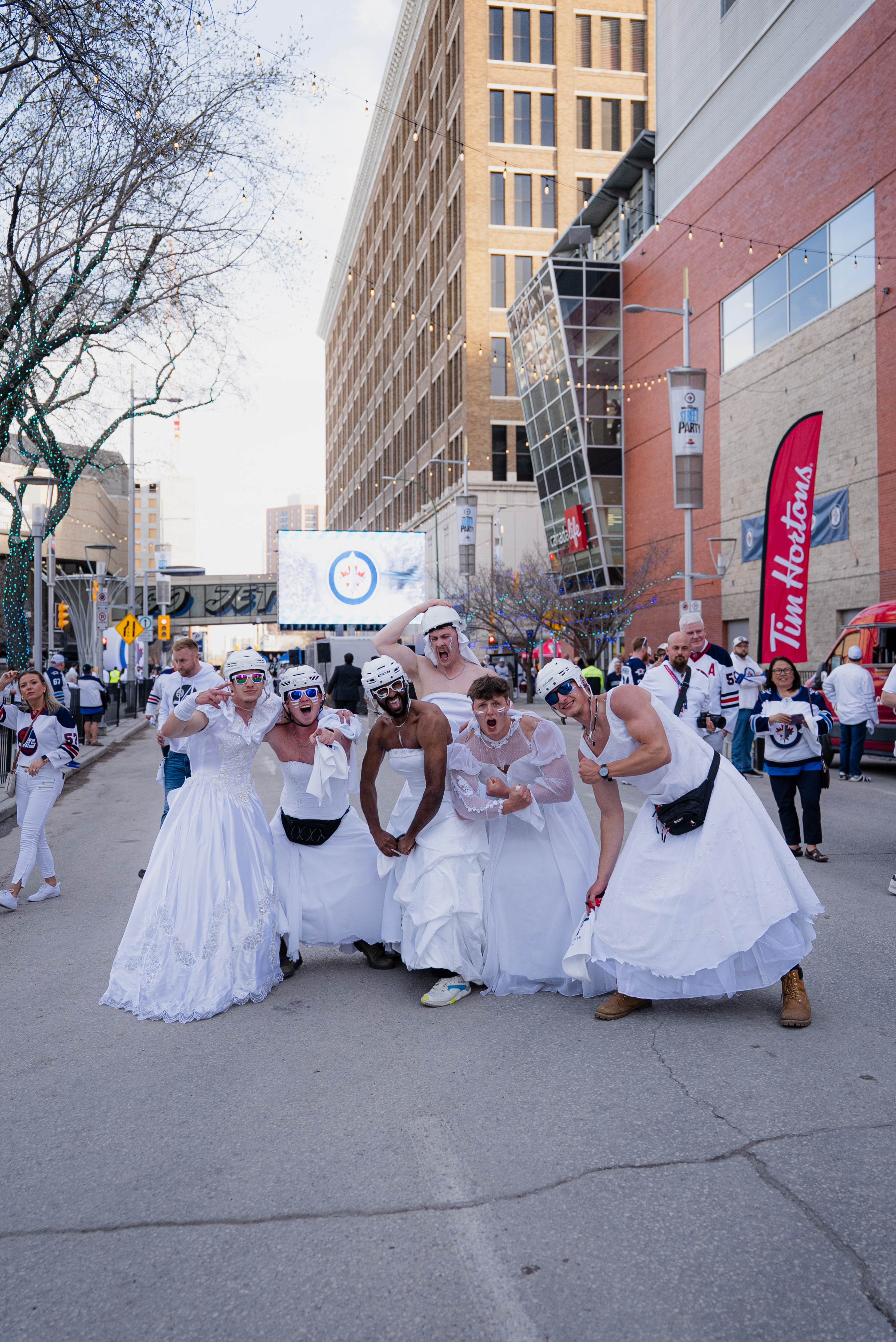 Six men posing in white wedding dresses and hockey helmets in downtown Winnipeg.