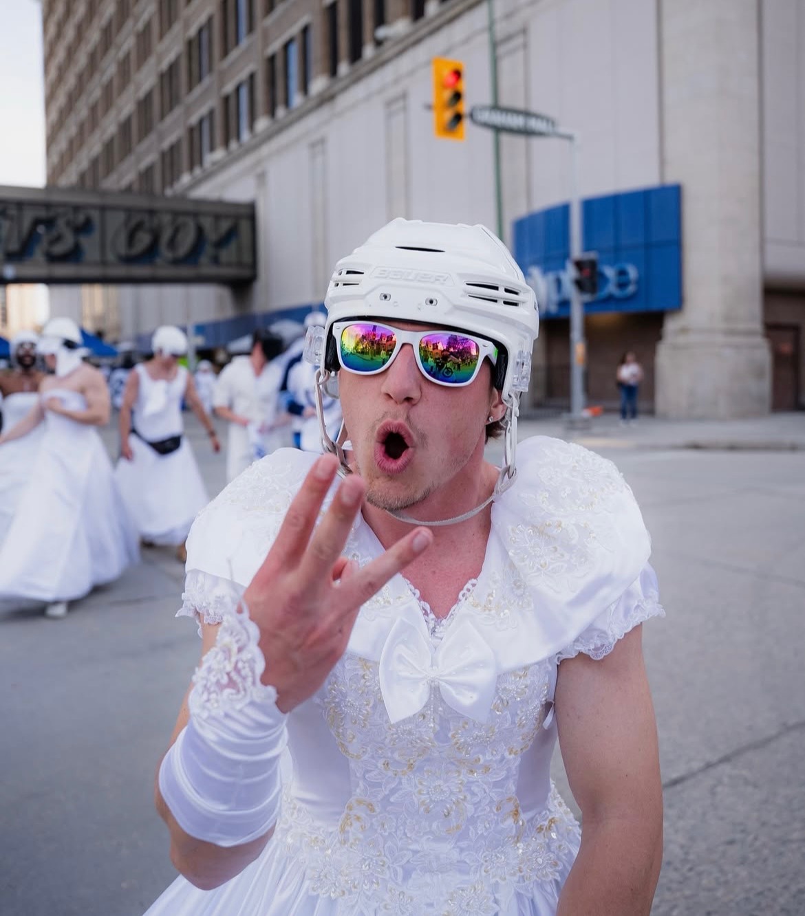 Man standing in downtown Winnipeg dressed in a white wedding dress, hockey helmet and sunglasses.