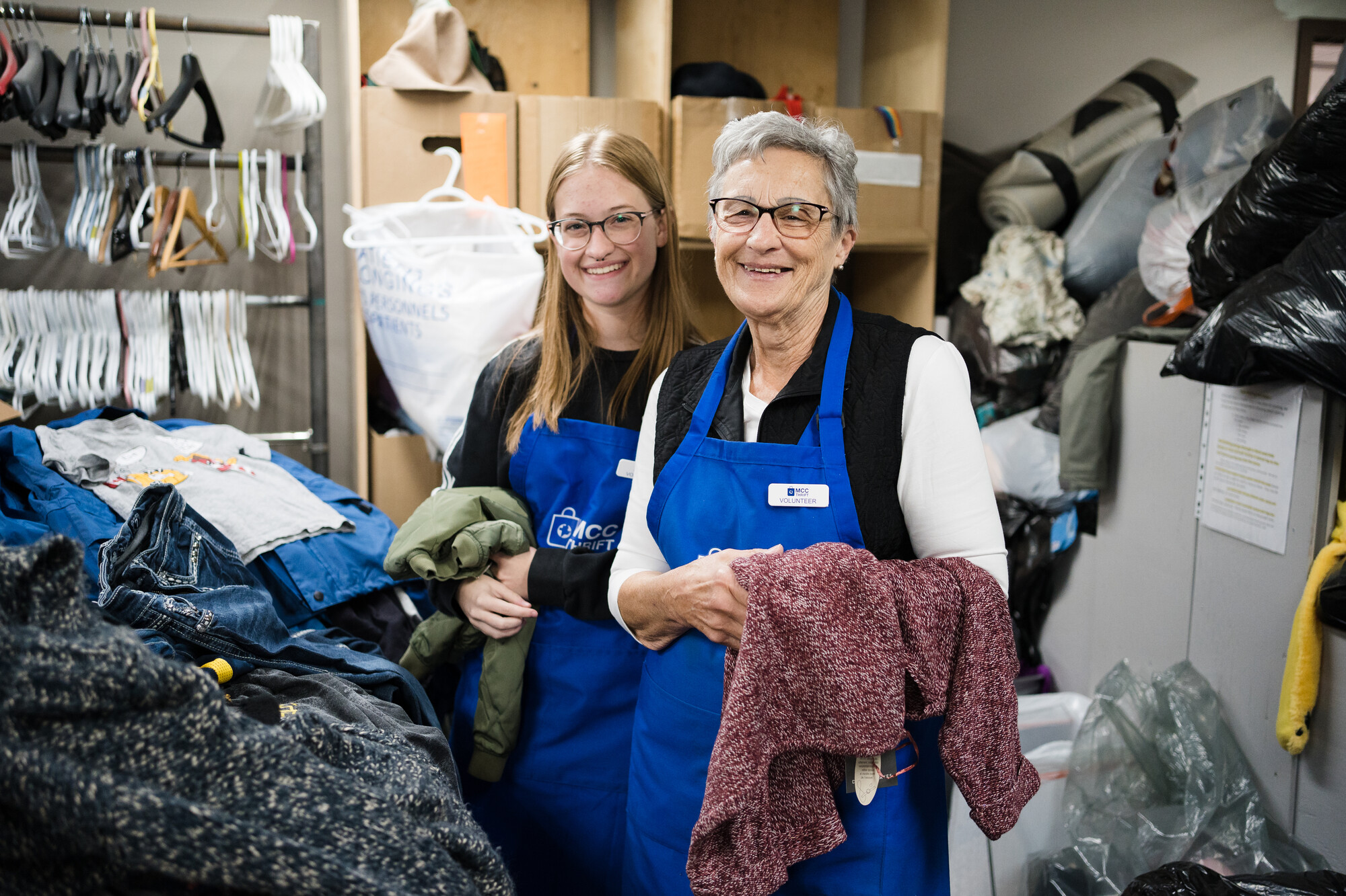 Two smiling women wearing blue aprons take a break sorting through clothing donations at an MCC Thrift Shop.
