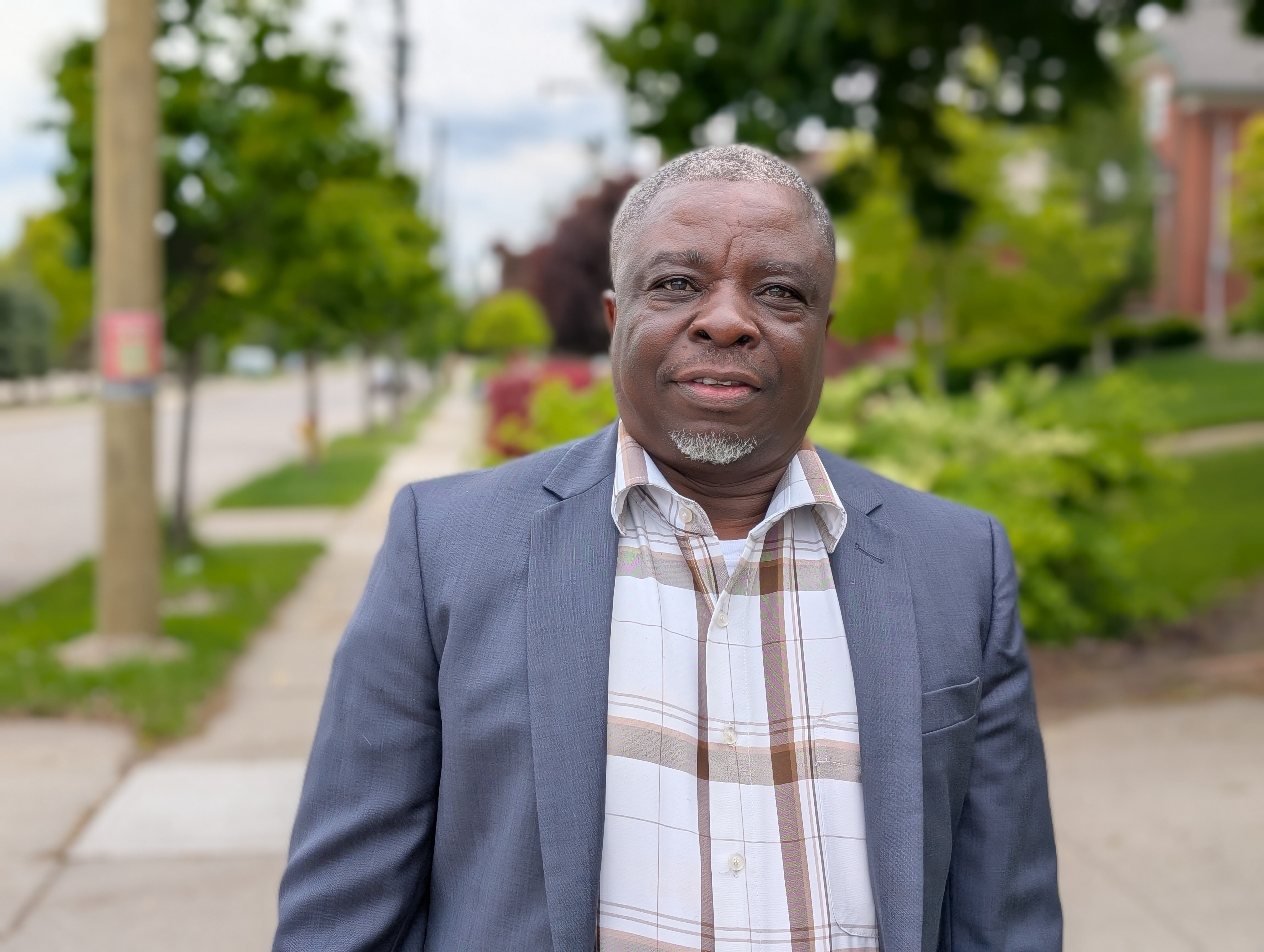 A middle-aged black man wearing a navy blazer and checked shirt look at the camera with a residential street in the background.