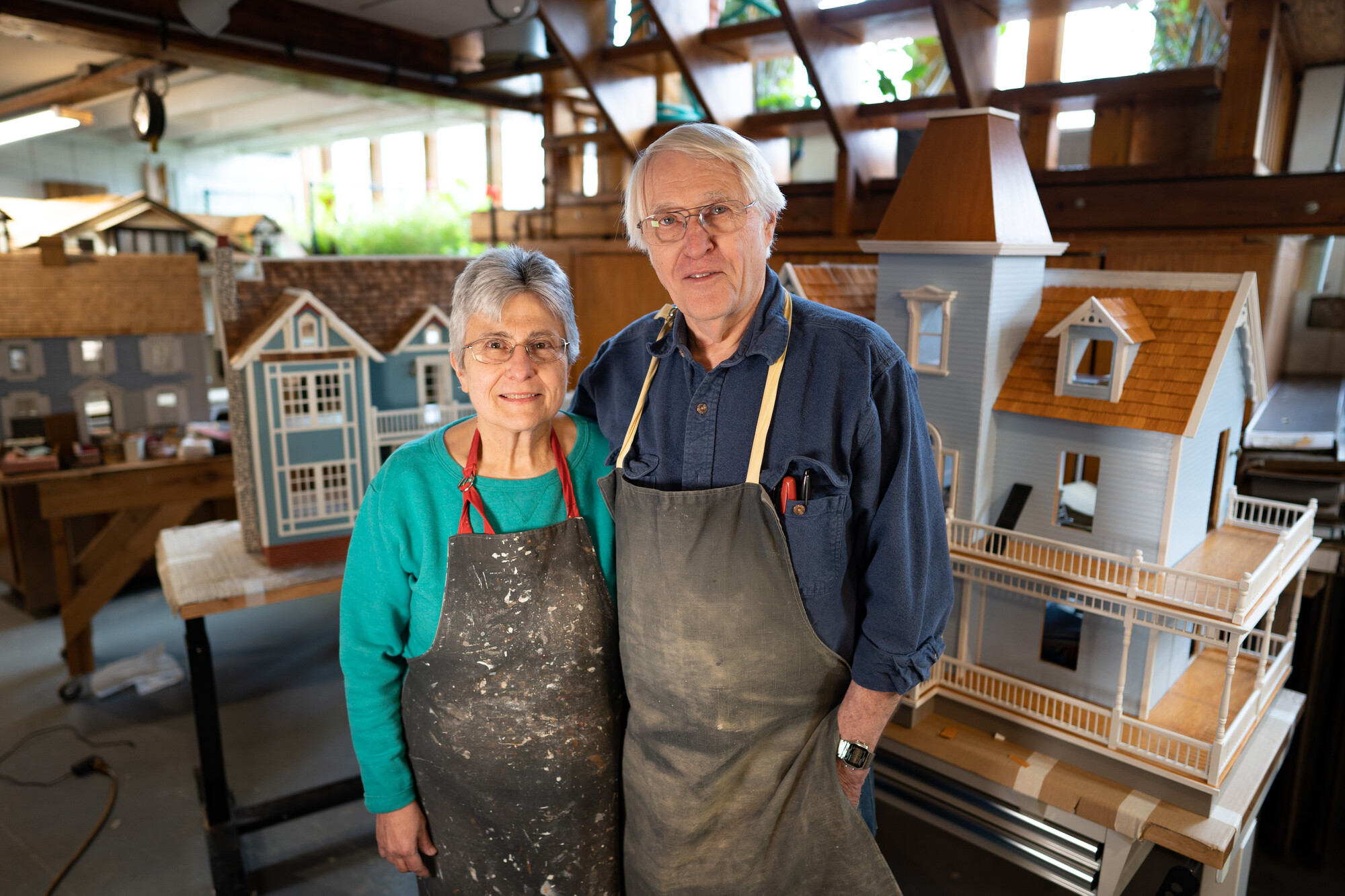 An older man and woman stand in a workshop featuring dollhouses. They are both wearing work aprons.