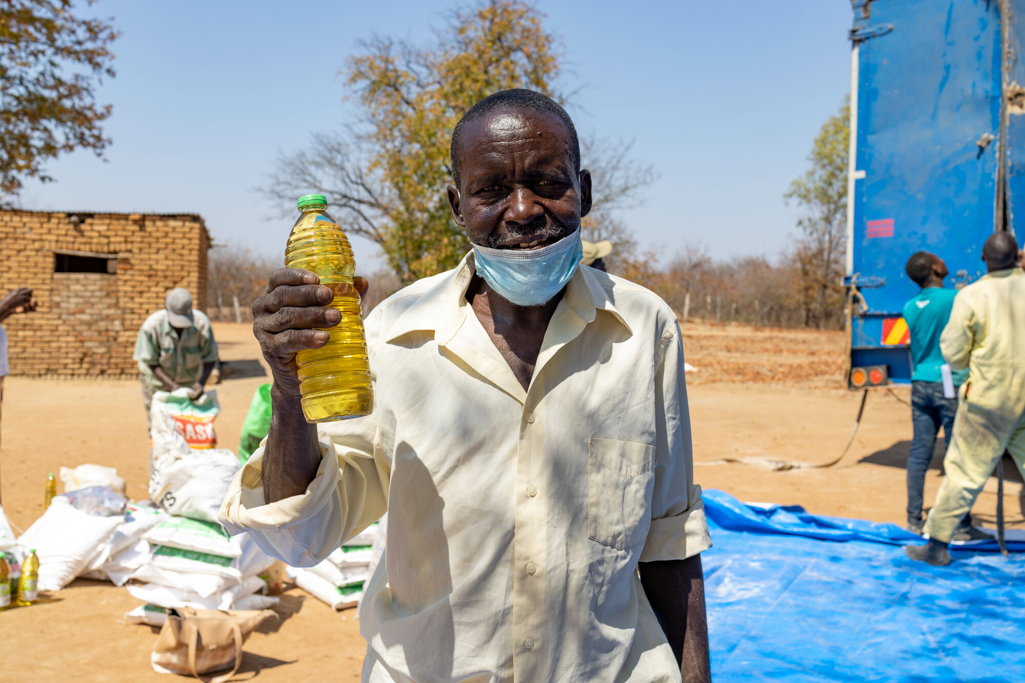 An older man with a face mask on this chin smiles for the camera and lifts up a plastic bottle of oil.