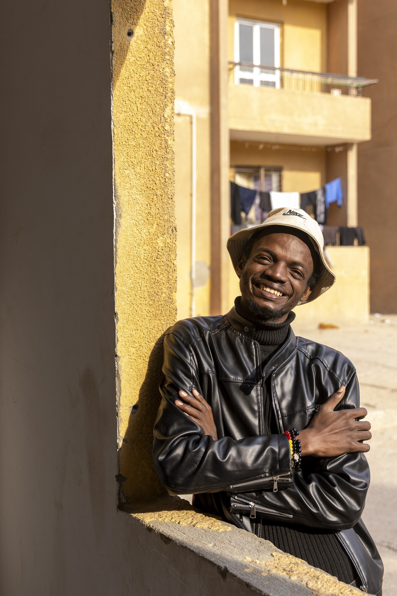 + A person in a leather jacket and hat stands by a wall, smiling. In the background, there are balconies with clothes hanging.