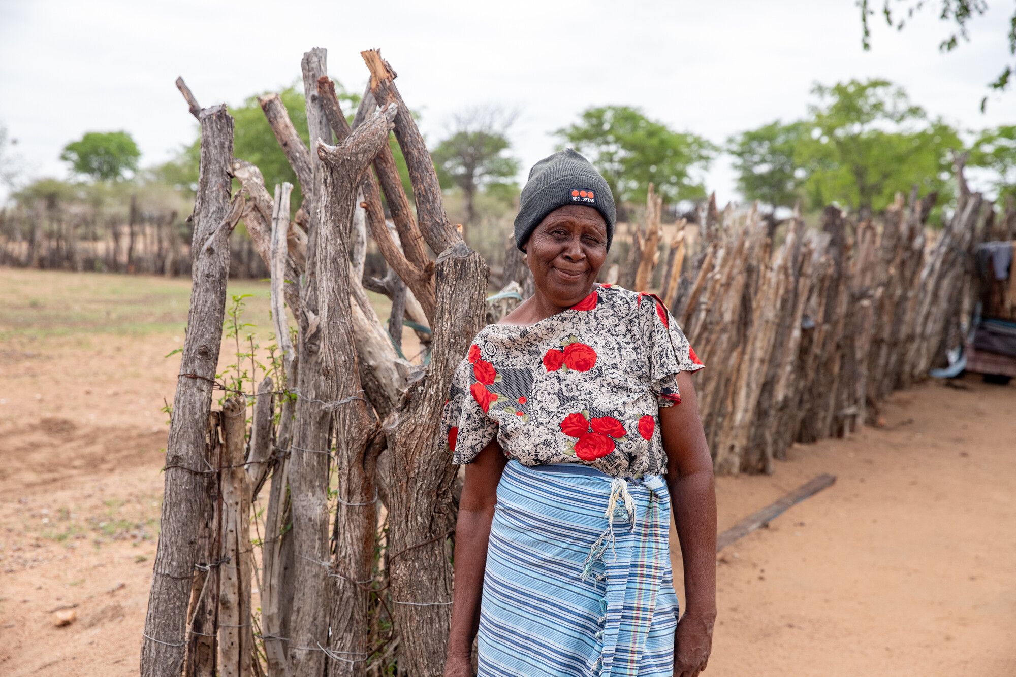 An elderly woman stands beside a wooden fence in a rural setting, wearing a patterned top and a head covering, with trees in the background.
