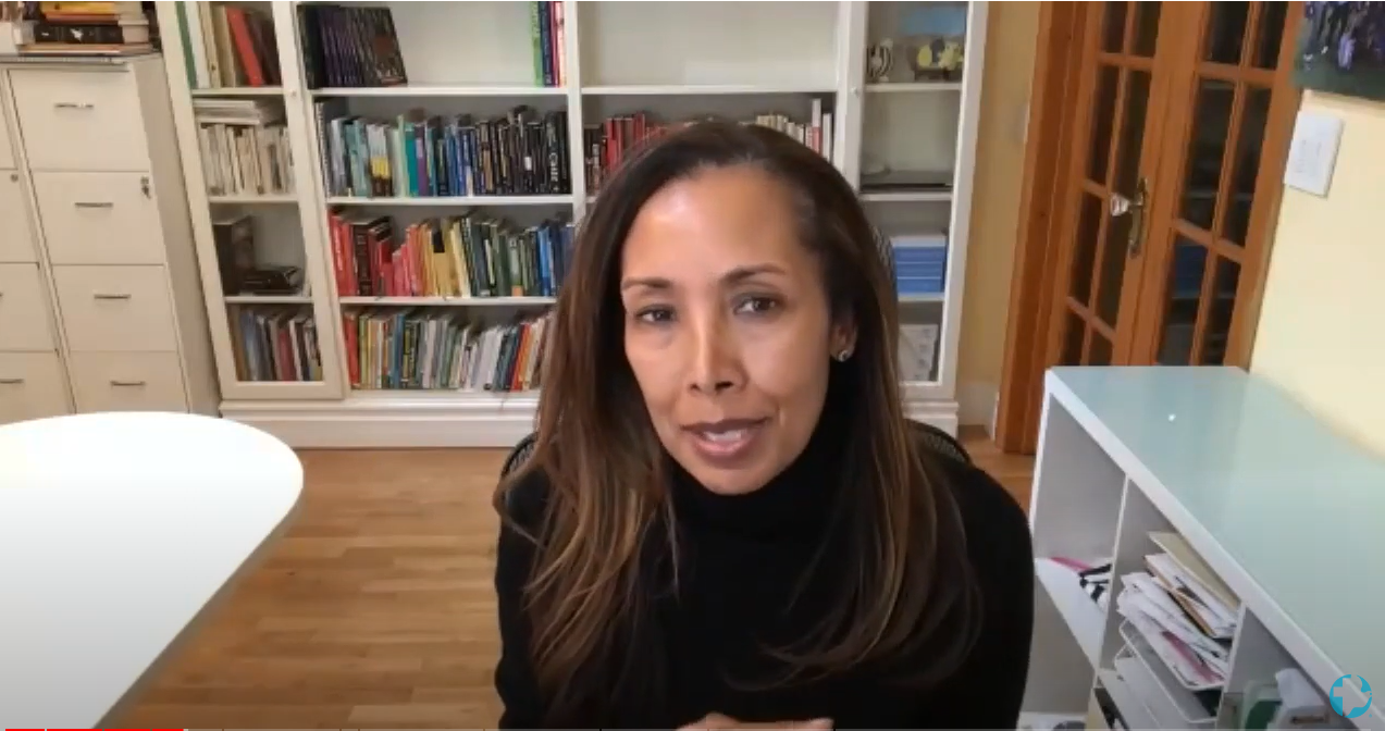 A woman with long hair and a black shirt speaks into the camera during a video call