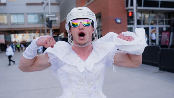 Man standing in downtown Winnipeg dressed in a white wedding dress, hockey helmet and sunglasses. More men in wedding dresses can be seen in the background.