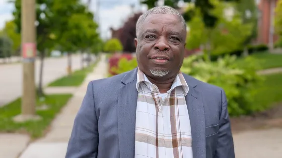 A middle-aged black man wearing a navy blazer and checked shirt look at the camera with a residential street in the background.