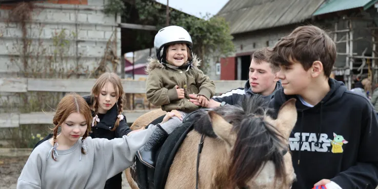 Three-year-old Vasylisa* sits calmly with assistance from volunteers during therapeutic horse riding for children who are displaced and affected by war.MCC, with partner Association of Mennonite B